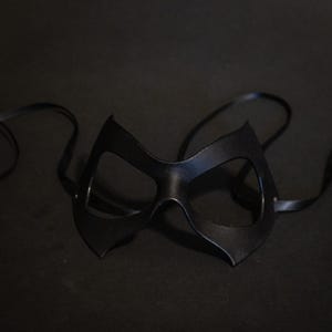 May include: A black eye mask with a sleek, cat-eye design. The mask is made of a smooth, dark material and features eye openings and attached black ribbons for securing. The mask is set against a dark background.