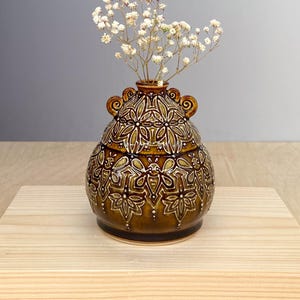 May include: A small, round, dark brown ceramic vase with a glossy finish and intricate, light-colored embossed floral patterns. The vase features two small, curled handles at the neck and contains a sprig of delicate, dried white baby's breath flowers.