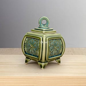 Può includere: Un barattolo quadrato in ceramica con coperchio, caratterizzato da una smaltatura verde salvia lucida e un manico circolare sulla parte superiore. I quattro lati presentano un pannello incassato con motivo floreale blu ottanio e oro.