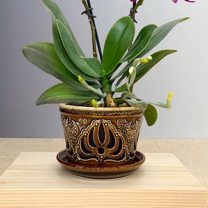 Könnte beinhalten: Eine leuchtend magentafarbene Orchidee mit zwei blühenden Stielen und grünen Blättern, eingetopft in einen braunen Keramikübertopf mit komplizierten Ausschnittmustern und passendem Untersetzer. Die Pflanze wird von zwei schwarzen Stäben gestützt.