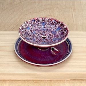 May include: A ceramic incense burner consisting of a deep red and blue glazed bowl with a detailed heart and floral pattern, featuring a central hole, resting on a matching circular red saucer with a small integrated holder for incense sticks.