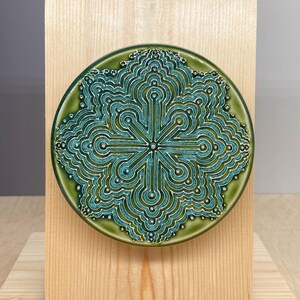 May include: A circular ceramic tile featuring a raised, intricate teal and green geometric mandala pattern with symmetrical lines and dots, mounted centrally on a rectangular light-colored wooden block stand with a visible wood grain texture.