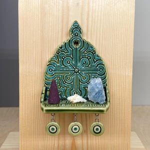 Op de afbeelding: Een decoratief turquoise keramisch rek met een kruisontwerp, gemonteerd op een houten plank. Het rek bevat een paarse wierookkegel, een witte schelp en een blauw kristal. Drie turquoise cirkels hangen eronder.