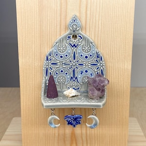 May include: A gray ceramic wall shelf with a blue and white floral design, mounted on a wooden board. The shelf holds a purple incense cone, a small shell, and a cluster of amethyst crystals. Decorative crescent moon and flower charms hang below.