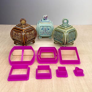May include: Three ceramic lidded jars with square bases and decorative handles, featuring brown, light blue, and green glazes with intricate patterns. Below them are seven bright pink plastic cookie cutters in various square and rectangular shapes.
