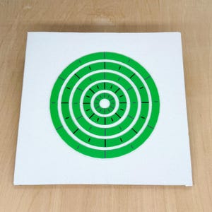 May include: A white square with a green target design. The target has concentric circles with black lines marking the intervals. The background is a light wood surface.