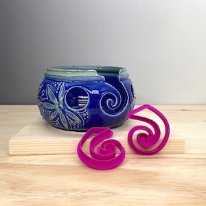 May include: A blue ceramic yarn bowl with a floral design and a swirl pattern. Two pink plastic yarn guides are on a wooden surface.