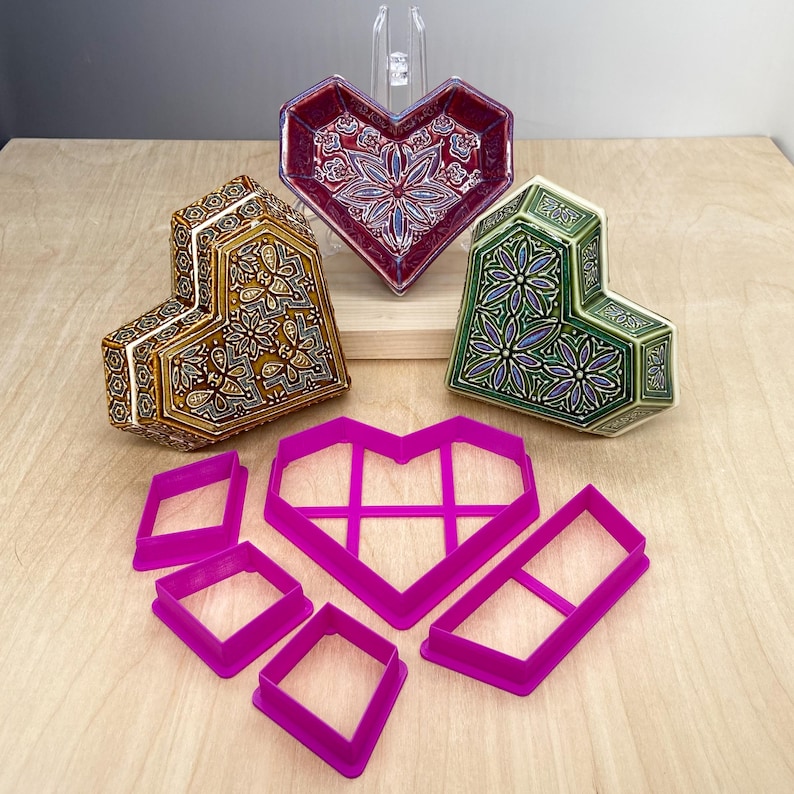 May include: Three heart-shaped ceramic boxes with intricate floral designs in shades of red, green, and gold.  The boxes are displayed on a wooden surface with five pink heart-shaped cookie cutters.