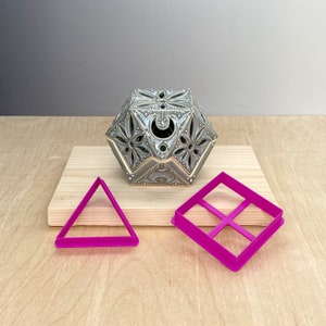 Op de afbeelding: Een grijs, geometrisch keramisch object met ingewikkelde bloemen en geometrische uitsparingen staat op een houten plank. Twee felroze plastic koekjessnijders, een driehoek en een vierkant met een kruis, liggen ervoor.