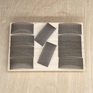 May include: A light-colored wooden board with several black mesh pieces arranged on top. The mesh pieces are rectangular and curved, with a grid-like pattern. The board is rectangular and has a natural wood grain.
