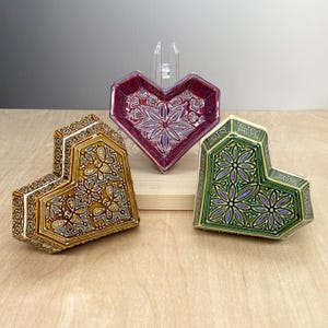 May include: Three ceramic heart-shaped boxes with intricate geometric designs. The boxes are in shades of green, gold, and purple.