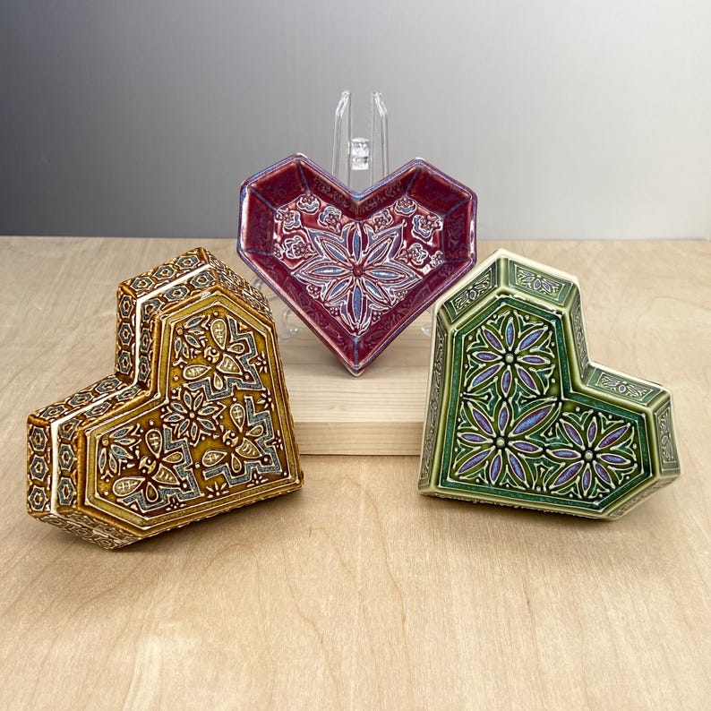 May include: Three ceramic heart-shaped boxes with intricate geometric designs. The boxes are in shades of green, gold, and purple.