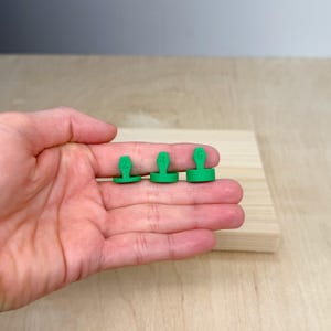 May include: Three small, green, numbered game pieces. The numbers 2, 4, and 6 are printed on the top of each piece. The pieces are held in a hand, and are resting on a light-colored wooden surface.