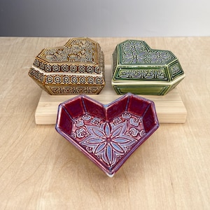 May include: Three heart-shaped ceramic boxes with intricate floral designs. The boxes are in shades of brown, green, and red.