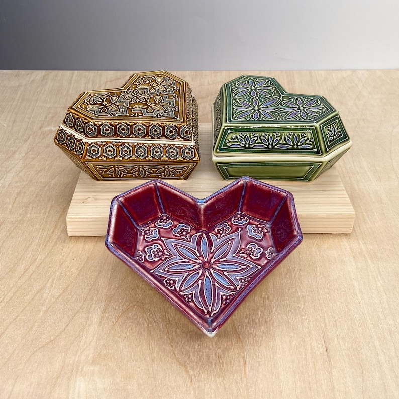 May include: Three heart-shaped ceramic boxes with intricate floral designs. The boxes are in shades of brown, green, and red.