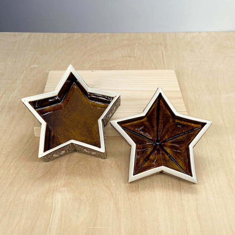 Puede incluir: Dos cuencos de cer&aacute;mica con forma de estrella. El interior de cada cuenco es de color marr&oacute;n intenso, mientras que los bordes son blancos. Los cuencos en forma de estrella tienen una superficie texturizada y est&aacute;n colocados sobre una tabla de madera. Los cuencos son decorativos.