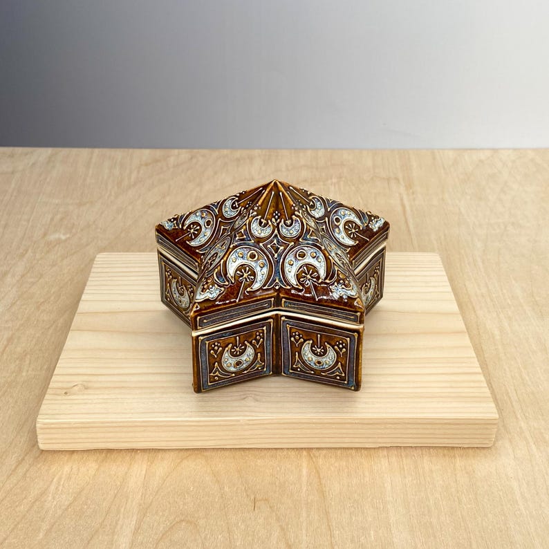 Puede incluir: Una caja decorativa en forma de estrella con tapa con bisagras. La caja es marr&oacute;n con intrincados dise&ntilde;os de luna creciente y flores blancas y plateadas. Se encuentra sobre una superficie de madera de color claro, mostrando su forma &uacute;nica y su detallada artesan&iacute;a.