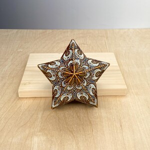 Puede incluir: Una estrella decorativa con un patr&oacute;n detallado en tonos marr&oacute;n, blanco y azul. La estrella tiene un dise&ntilde;o central en relieve y est&aacute; colocada sobre una superficie de madera clara. El dise&ntilde;o de la estrella es intrincado y ornamentado.