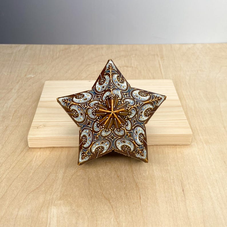Puede incluir: Una estrella decorativa con un patr&oacute;n detallado en tonos marr&oacute;n, blanco y azul. La estrella tiene un dise&ntilde;o central en relieve y est&aacute; colocada sobre una superficie de madera clara. El dise&ntilde;o de la estrella es intrincado y ornamentado.