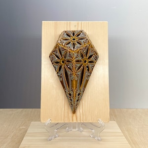 Puede incluir: Un colgante de pared de cerámica marrón en forma de diamante con un patrón floral decorativo. El diamante está montado en una placa de madera con un soporte.