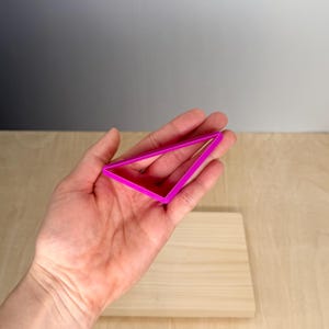 Puede incluir: Un cortador de galletas triangular de color rosa brillante sostenido en una mano. El cortador es de pl&aacute;stico y tiene un dise&ntilde;o geom&eacute;trico simple. El fondo es una superficie de madera clara.