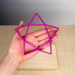 Puede incluir: Un cortador de galletas en forma de estrella magenta brillante. El cortador se sostiene sobre una superficie de madera de color claro. La estrella tiene un dise&ntilde;o de cinco puntas con bordes rectos y una forma geom&eacute;trica. El fondo es un degradado de gris y blanco.