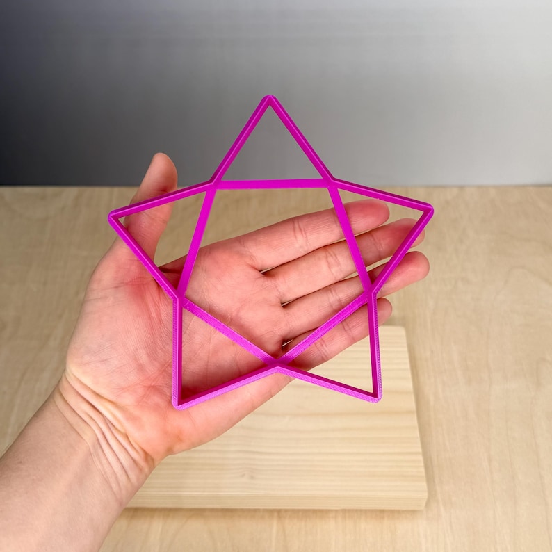 Puede incluir: Un cortador de galletas en forma de estrella magenta brillante. El cortador se sostiene sobre una superficie de madera de color claro. La estrella tiene un dise&ntilde;o de cinco puntas con bordes rectos y una forma geom&eacute;trica. El fondo es un degradado de gris y blanco.