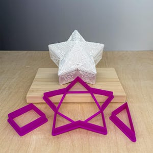 May include: A white, star-shaped decorative box with ornate embossed detailing sits on a wooden board. Below are three magenta-colored cookie cutters: a square, a star, and a triangle. The background is a neutral gray.
