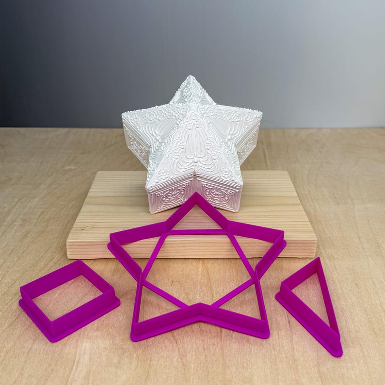 Puede incluir: Una caja decorativa blanca en forma de estrella con detalles en relieve ornamentales se encuentra sobre una tabla de madera. Debajo, tres cortadores de galletas magenta: un cuadrado, una estrella y un tri&aacute;ngulo. El fondo es gris neutro.