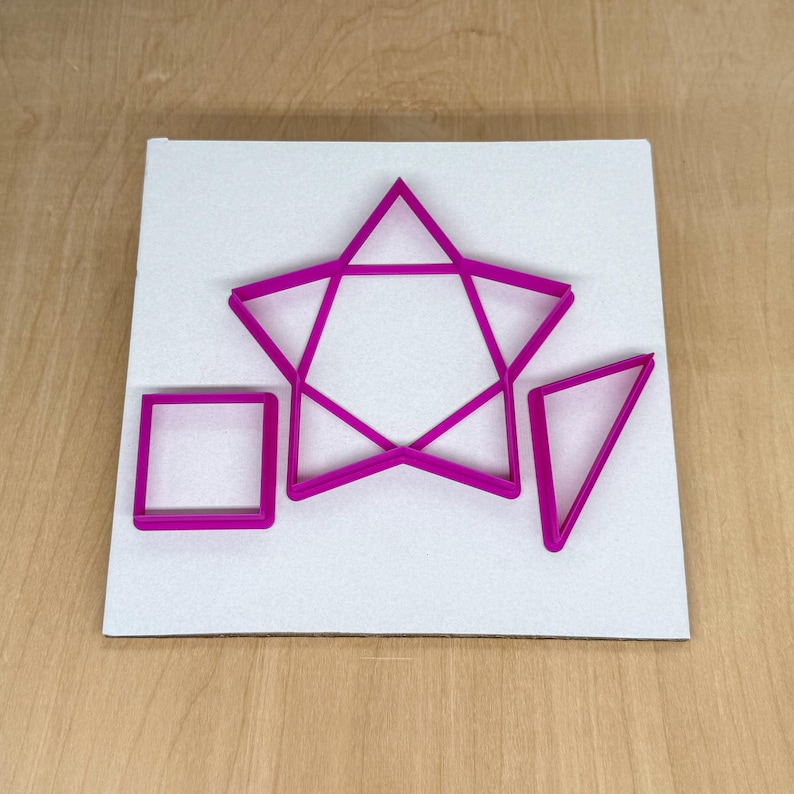Puede incluir: Un conjunto de cortadores de galletas con formas geom&eacute;tricas de color rosa brillante, que incluyen un cuadrado, un tri&aacute;ngulo y una estrella de cinco puntas, dispuestos sobre una superficie blanca. Las formas son de pl&aacute;stico y est&aacute;n dise&ntilde;adas para hornear o manualidades.