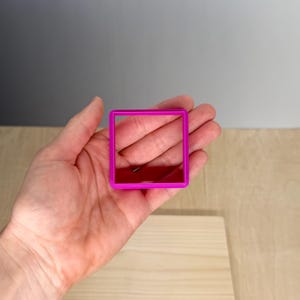 Puede incluir: Cortador de galletas cuadrado rosa brillante sostenido en una mano. El cortador es de pl&aacute;stico y tiene un dise&ntilde;o geom&eacute;trico simple. El fondo es una superficie de madera clara y una pared gris.
