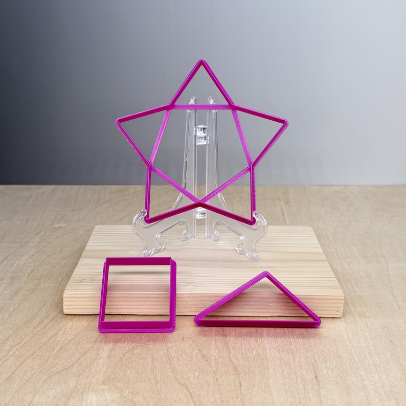 Puede incluir: Un conjunto de tres cortadores de galletas geom&eacute;tricos de color magenta. Una forma de estrella se muestra en un soporte transparente, con un cuadrado y un tri&aacute;ngulo planos sobre una superficie de madera. Las formas son para hornear.
