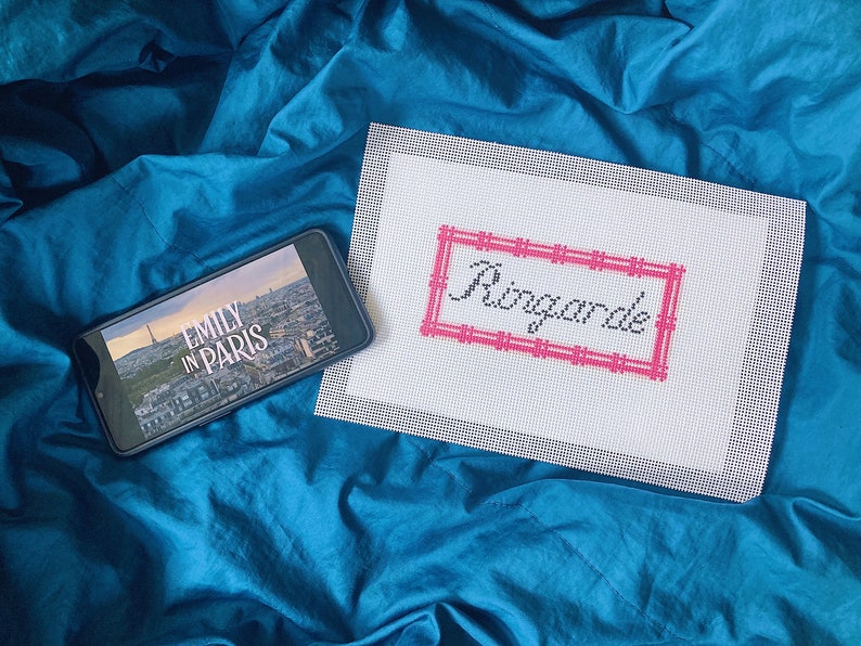 May include: A smartphone displaying the Netflix show "Emily in Paris" on a blue bedspread next to a white canvas with the words "Ringarde" embroidered in black thread with a pink border.