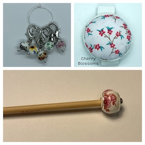 May include: A set of stitch markers with floral designs, a pincushion with a white background and pink cherry blossom pattern, and a bamboo knitting needle with a floral bead. The pincushion has the text "Cherry Blossoms" printed on it.