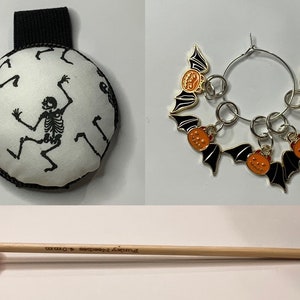 May include: Halloween themed crochet supplies including a round stitch marker with a skeleton design, a set of four stitch markers with bat and pumpkin designs, and a crochet hook with a pumpkin-shaped handle.