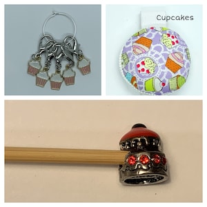 May include: Set of five cupcake-shaped stitch markers with silver rings and a silver wire loop. The stitch markers are pink and white with a silver cupcake liner. The stitch markers are hanging from a silver wire loop. A close-up of a silver cupcake stitch marker with red and clear rhinestones. The stitch marker is on a wooden knitting needle. A round pincushion with a purple background and a white lace pattern. The pincushion is covered in a fabric with a cupcake pattern. The pincushion has the word "Cupcakes" printed on a white tag.