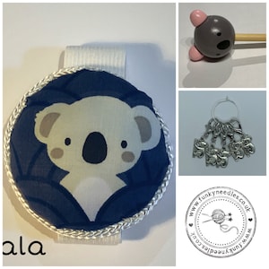 May include: A blue and white koala stitch marker with a white elastic band. The stitch marker is round and has a white border. The koala is in the center of the marker and has a cute face. The text "ala" is visible in the bottom left corner of the image.