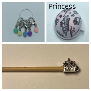 May include: A set of five pastel-colored heart-shaped stitch markers with silver rings, a silver pincushion with a white border and a pink fabric with a princess design, and a wooden knitting needle with a silver castle-shaped end cap.