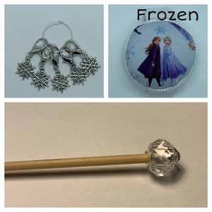 May include: A set of five silver snowflake stitch markers with a silver ring and a silver clasp. The stitch markers are hanging from the ring. The stitch markers are on a white background. The image also shows a round pincushion with a Frozen theme. The pincushion is white with a blue and purple design featuring Anna and Elsa from the movie Frozen. The text "Frozen" is printed in black above the pincushion. The image also shows a wooden knitting needle with a clear crystal bead on the end.