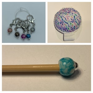 May include: A collection of knitting accessories. Includes a set of silver stitch markers with colorful beads, a pin cushion with a white ribbon, and a single bead on a wooden knitting needle. The bead is blue and white.