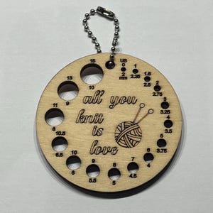 May include: A circular wooden knitting needle gauge with a keychain. The gauge has 10 holes for different needle sizes, labelled in both US and mm sizes. The gauge also has the text "all you knit is love" and a small illustration of yarn and knitting needles.