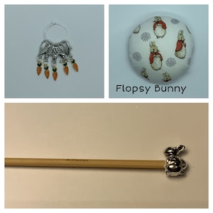 May include: A set of knitting stitch markers featuring a carrot design. The markers are silver with orange and green accents. The set also includes a round needle minder with a Peter Rabbit fabric design. The needle minder features a white background with a repeating pattern of gray dots and Peter Rabbit characters wearing red jackets. The text "Flopsy Bunny" is printed below the needle minder. A wooden knitting needle with a silver rabbit charm is also included.
