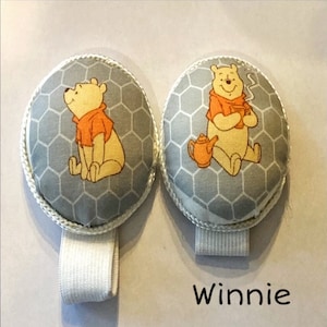 May include: Two gray and white fabric Winnie the Pooh themed baby stroller straps with a honeycomb pattern and Winnie the Pooh images. The straps are attached to a white elastic band. The text "Winnie" is written below the straps.