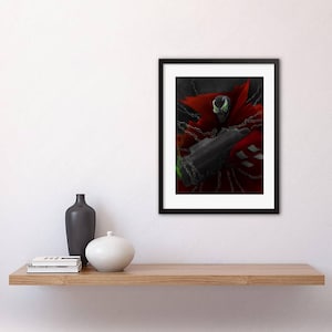 Può includere: Una stampa incorniciata in nero di un personaggio di fumetti, Spawn, con un mantello rosso e catene, che tiene una pistola. Il personaggio ha gli occhi verdi e un'espressione minacciosa.
