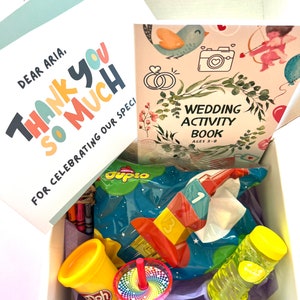May include: A gift box filled with various children's toys and activities, including a book titled "Wedding Activity Book Ages 3-8", a bag of Duplo blocks, a container of Play-Doh, a spinning top, and a bottle of bubbles. The box also includes a card that says "Dear Aria, Thank You So Much For Celebrating Our Special Day."