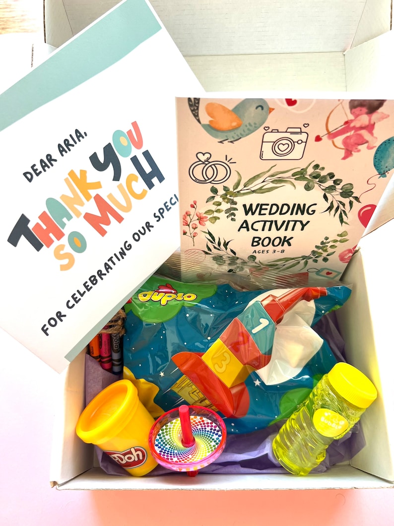 May include: A gift box filled with various children's toys and activities, including a book titled "Wedding Activity Book Ages 3-8", a bag of Duplo blocks, a container of Play-Doh, a spinning top, and a bottle of bubbles. The box also includes a card that says "Dear Aria, Thank You So Much For Celebrating Our Special Day."