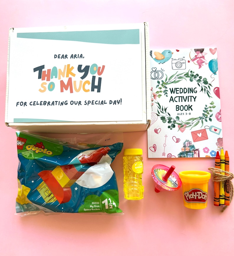 May include: A white box with the text "Dear Aria. Thank you so much for celebrating our special day!"  The box contains a variety of children's toys, including a Lego Duplo set, a bubble bottle, a spinning top, a container of Play-Doh, and a set of crayons.