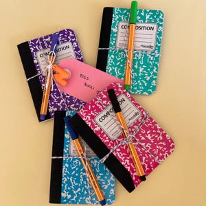 May include: Several composition notebooks in shades of purple, teal, pink, and blue, each with a black spine and a patterned cover. The notebooks are tied with twine and include pens in various colours, plus a pink tag that says "Miss Nikki."
