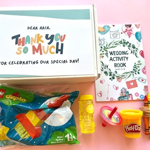 May include: A white box with the text "Dear Aria. Thank you so much for celebrating our special day!"  The box contains a variety of toys, including a bag of Lego Duplo blocks, a bubble wand, a spinning top, a container of Play-Doh, and a set of crayons.