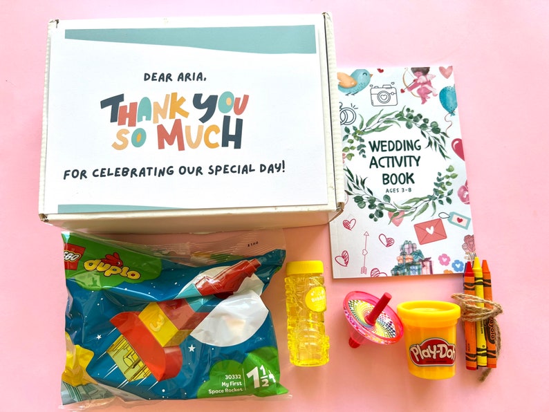 May include: A white box with the text "Dear Aria. Thank you so much for celebrating our special day!"  The box contains a variety of toys, including a bag of Lego Duplo blocks, a bubble wand, a spinning top, a container of Play-Doh, and a set of crayons.